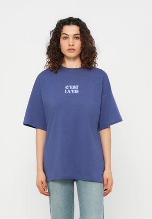 Νεαρό άτομο με σγουρά μαλλιά που φοράει φαρδιά μπλε μπλούζα τυπωμένη με τη φράση "C'EST LA VIE" και ανοιχτόχρωμο τζιν, μπροστά σε απλό φόντο.