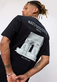 Homme avec des dreadlocks portant un T-shirt noir avec un croquis blanc de l'Arc de Triomphe et le mot "RENAISSANCE" dans le dos.