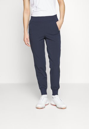 Person wearing navy jogger pants with elastic cuffs and white sneakers, standing with one hand in pocket against a plain background.