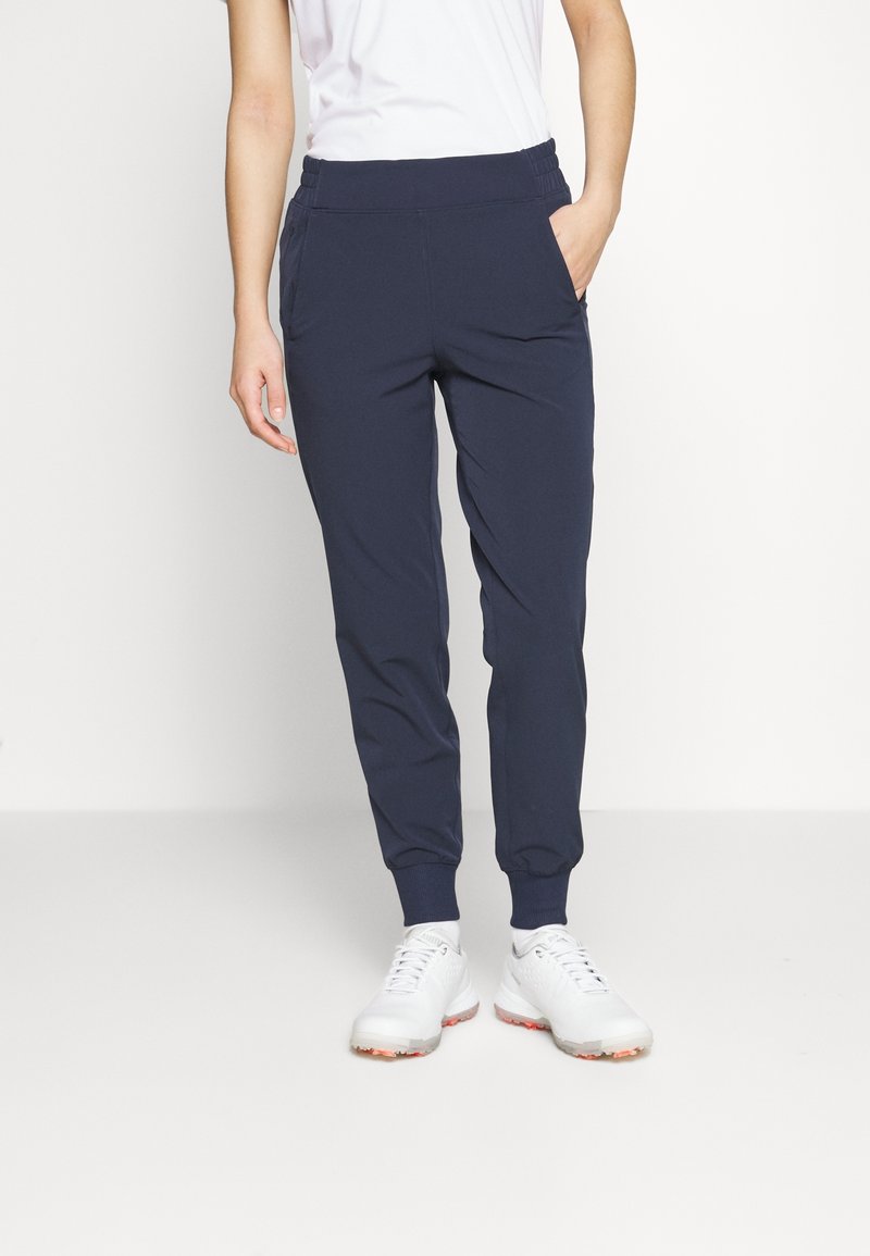 Personne portant un pantalon de jogging bleu marine avec des poignets élastiques et des baskets blanches, debout avec une main dans la poche devant un fond uni.
