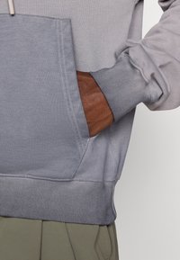 Siva dukserica s kapuljačom koja ima kenguruhu džep s smešnim kožnim detaljem i rebrastim manžetama i donjim rubom; glatka tekstura tkanine i opušten kroj.