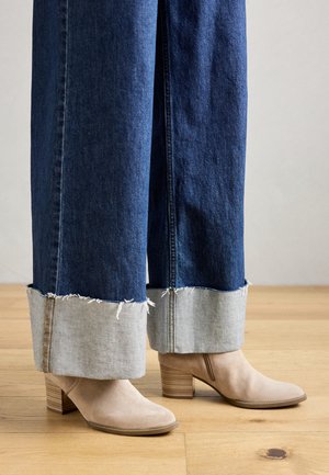 Beige suède enkellaarsjes met gestapelde houten hakken, gecombineerd met wijde donkerblauwe spijkerbroeken met een rafelige zoom en opgerolde cuffs.