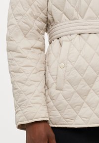 Beige gesteppte Jacke mit einem taillierten Gürtel, die über eine Rautenmustersteppung und eine seitliche Tasche mit Druckknopfverschluss verfügt. Glatte Textur.