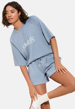 Lyseblåt sæt med afslappet loungebeklædning bestående af en løs, kortærmet sweatshirt med trykt logo og matchende shorts med snøre. Blødt stof, afslappet design.