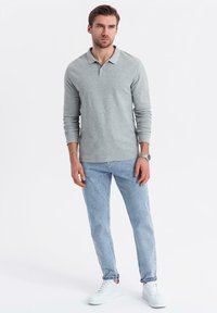 Grå långärmad polo skjorta, ljusblå jeans och vita sneakers bärs av en stående manlig figur, som visar en avslappnad outfit.