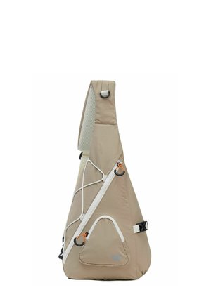 Mochila beige tipo bandolera con correa acolchada, bolsillo frontal con cremallera, hebillas laterales y detalle con cordón elástico sobre un fondo blanco.