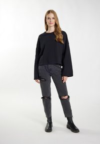 Schwarzer, kurzer Sweatshirt mit langen Ärmeln, kombiniert mit grauen, zerrissenen Jeans. Das Modell trägt schwarze Stiefeletten. Einfaches, lässiges Design und Passform.