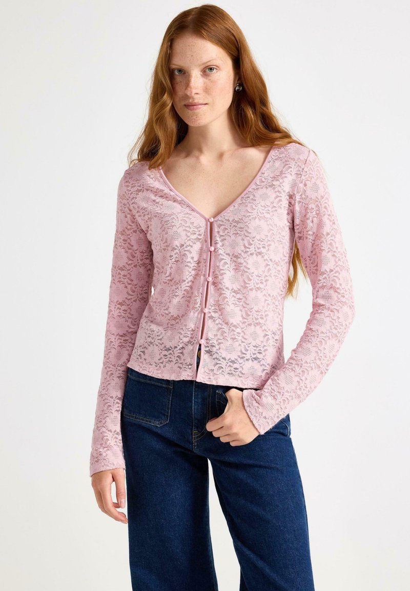Ung kvinde med langt rødt hår iført en lyserød blonde cardigan med knapper og mørkeblå jeans, stående med den ene hånd i lommen.