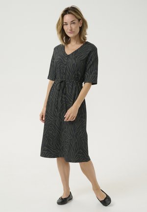 Robe noire avec un motif zèbre vert, décolleté en V, manches courtes, taille cintrée avec un cordon de serrage et ourlet à hauteur du genou. Portée avec des ballerines noires.
