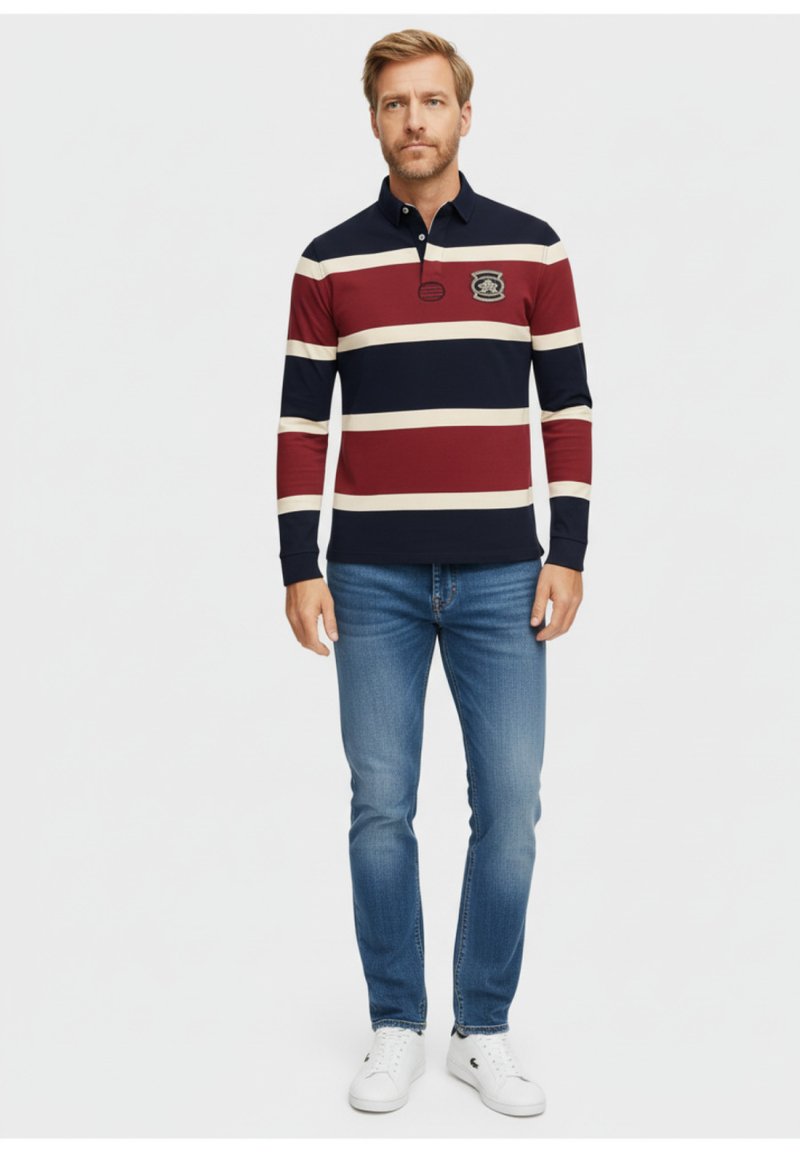 Camisa polo de manga larga con rayas en navy, burdeos y crema, con un cuello y un parche bordado en el pecho. Lleva puesta con jeans claros.