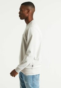 Sudadera gris claro con puños acanalados, escote redondo y aberturas laterales. Combinada con jeans desgastados de color azul claro. La textura parece suave.