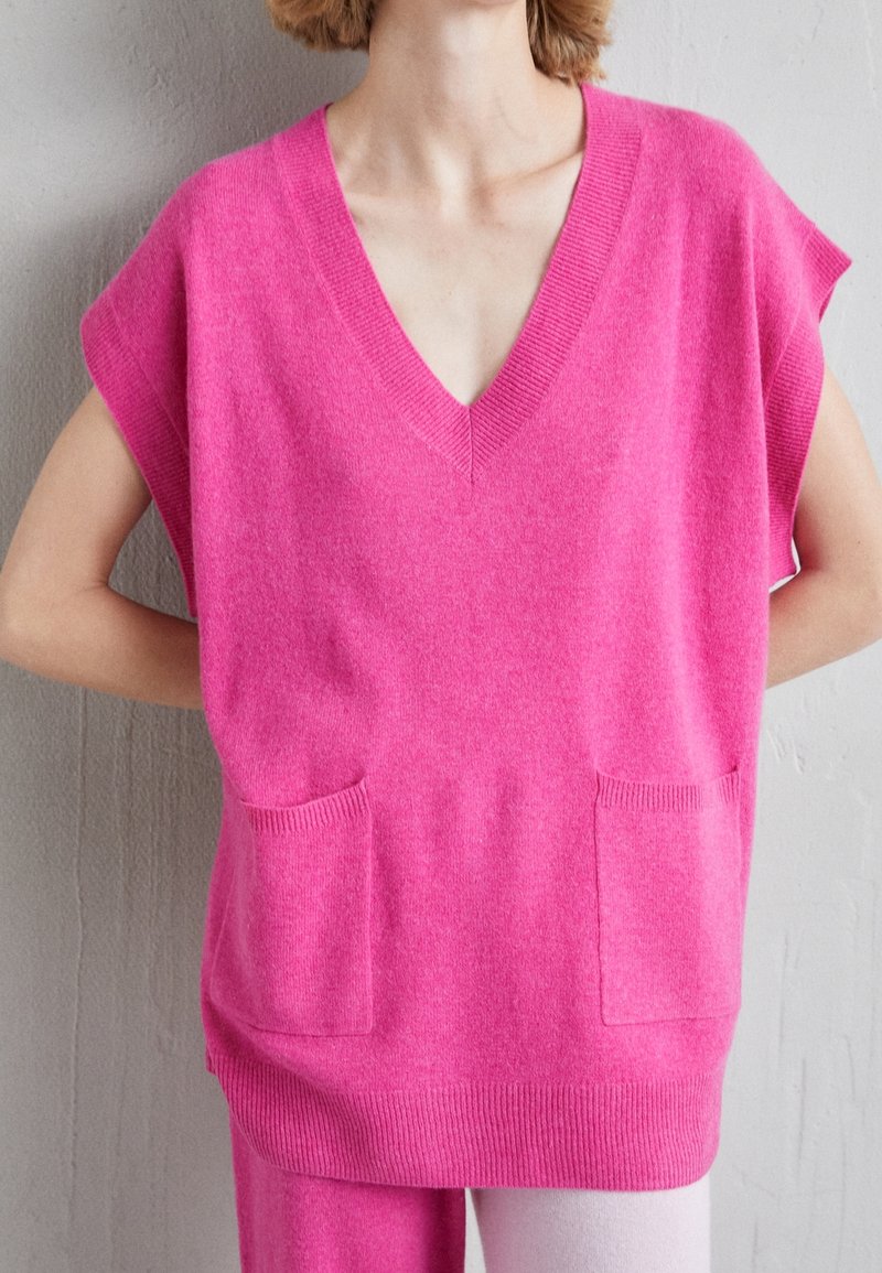 Femme portant un gilet en maille sans manches rose vif avec un col en V et des poches avant, debout devant un mur uni gris clair.