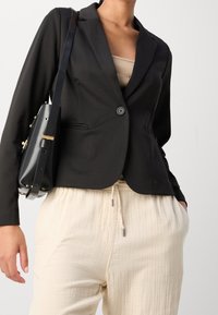 Černý blazer s klopami a jedním knoflíkem, kombinovaný se světle béžovými kalhotami. Malá černá kabelka přes rameno se zlatými akcenty.