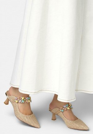 Pieds portant des mules tissées beige à bout pointu avec des talons bas et des brides ornées de pierres colorées, associées à une longue jupe blanche.