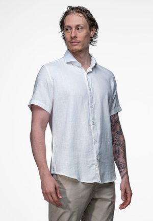 Man met tatoeages op zijn rechterarm draagt een wit overhemd met korte mouwen en knopen en beige broek, kijkt naar links tegen een effen achtergrond.