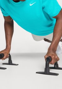 Czarne poręcze do pompek na jasnym tle, z osobą w turkusowej koszulce Nike wykonującą pompkę. Poręcze mają gumowe uchwyty i solidną podstawę.