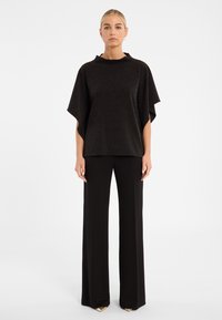 Schwarzes, glitzerndes Poncho-Style-Oberteil mit hohem Ausschnitt und weiten Ärmeln, kombiniert mit eng anliegenden schwarzen Hosen und spitzen, goldenen Schuhen.