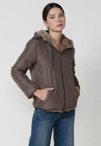 Rjava podložena jakna z zadrgo in kapuco, ki ima mehak umetni krznen rob. Vertikalne šive in stranske žepe povečujejo funkcionalnost.