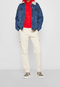 Džínová bunda s kožešinovým límcem, červená mikina, krémové cargo kalhoty a černobílé kostkované slip-on boty. Neformální vrstvený outfit.