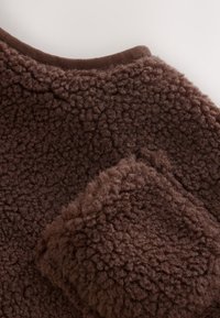 Gros plan d’un tissu en polaire marron texturé montrant une surface douce en peluche et un détail de poche près du bord du col.