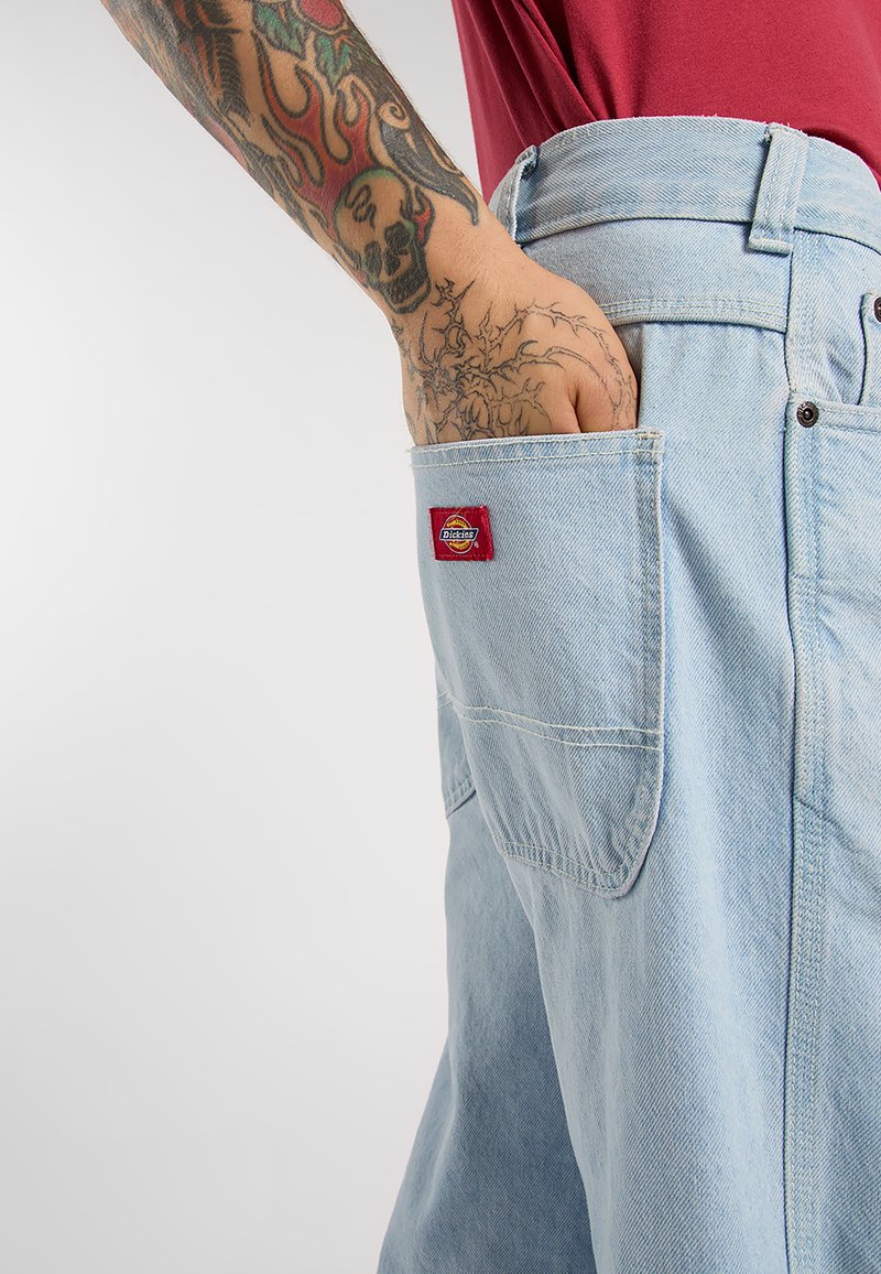 Getatoeëerde arm met schedel en vlammen rust in de achterzak van lichtblauwe Dickies-jeans, gecombineerd met een rood shirt.