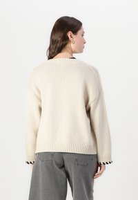 Beige gestrickter Pullover mit lockerer Passform, geripptem Saum und Bündchen. Getragen über grauen hoch taillierten Hosen.