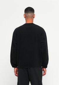 Mand med kort hår iført en løs sort sweatshirt og sorte cargo-bukser, stående vendt mod en simpel hvid baggrund.
