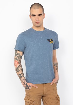 T-shirt blu a collo rotondo con una texture sbiadita, caratterizzato da un logo alato nero e giallo sul petto. Maniche corte.