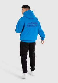 Felpa blu con vestibilità oversize, con la scritta "no limits just Sport" in blu scuro sulla schiena. Indossata con pantaloni della tuta neri e sneakers bianche.