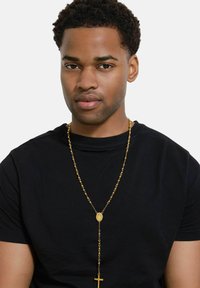 Collana in oro con catena e medaglione ovale con pendente a croce, caratterizzata da un design con perline e una superficie liscia, indossata sopra una t-shirt nera.