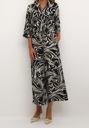 Kvinde iført en sort og hvid abstrakt mønstret buksedragt med vide ben og trekvartærmer, kombineret med cremefarvede højhælede mules.