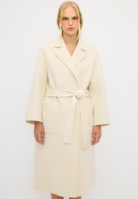 Beige Wollmantel mit taillierter Gürtellinie, Reverskragen, weiten Ärmeln und zwei großen vorderen Taschen. Glatte Textur und knielanges Design.