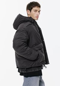 Jachetă puffer neagră cu glugă, prevăzută cu închidere cu fermoar și capse, șnur elastic la tiv și buzunare laterale. Textură netedă.