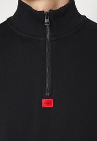 Gros plan d’un pull noir à fermeture éclair quart avec un écusson carré rouge portant le logo "HUGO" sous la fermeture éclair sur la poitrine.