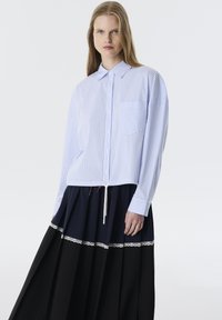 Camisa de rayas azul y blanca con cuello y un bolsillo en el frente, combinada con una falda larga y plisada que presenta acentos en negro, azul marino y encaje.