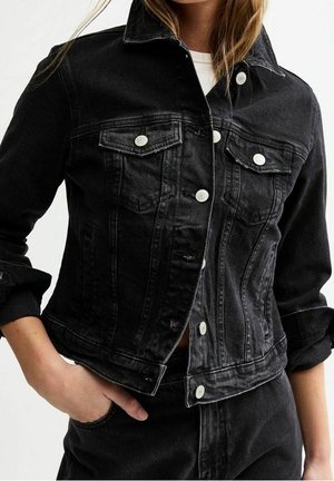 Nainen pukeutuneena mustaan, istuvaan farkkutakkiin, jossa on hopeiset napit, kaksi rintataskua ja käärityt hihat, yhdistettynä mustiin housuihin.