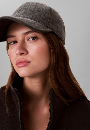 Grijze pet met een gebogen klep, gestructureerde stof en een subtiel patroon. Model draagt een donkere fleecevest met rits eronder.