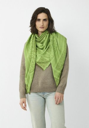 Grüner, gemusterter Schal aus weichem Stoff, der komplizierte Designs und Fransenkanten aufweist, über einem beigen Pullover getragen.