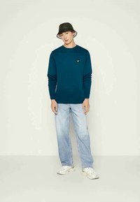 Sweatshirt teal com mangas longas apresentando um pequeno logótipo amarelo, combinado com jeans azul claro e ténis brancos. Chapéu de balde preto.