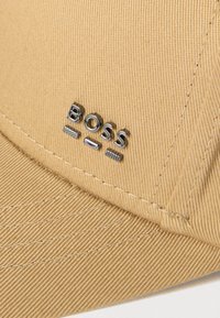 Czapka bejsbolowa w kolorze khaki o fakturowanej powierzchni, z naszytym na froncie srebrnym logo "BOSS". Posiada zakrzywiony daszek oraz regulowane zapięcie z tyłu.