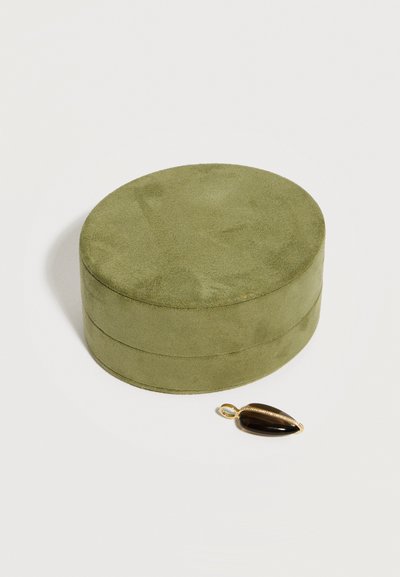 Στρογγυλό κουτί κοσμημάτων από σουέτ ελιάς με ένα χρυσό και μαύρο μενταγιόν σε σχήμα σταγόνας δίπλα του, πάνω σε λευκή επιφάνεια.