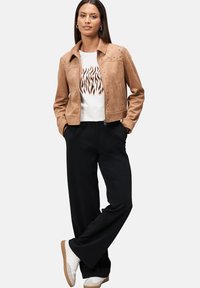Tan ruskindsjakke med nitter, hvid grafisk t-shirt og sorte vide bukser, parret med hvide sneakers på en neutral baggrund.