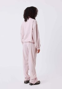 Persona con cabello rizado que lleva un chándal holgado de color rosa pálido y zapatillas negras gruesas, de pie contra un fondo claro y liso.