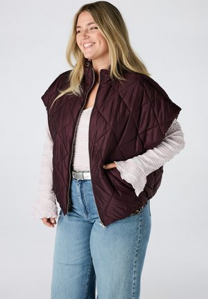 Lachende vrouw die een donkerpaarse doorgestikte bodywarmer draagt over een lichtroze longsleeve en blauwe spijkerbroek met een zilveren riem, staand tegen een effen achtergrond.
