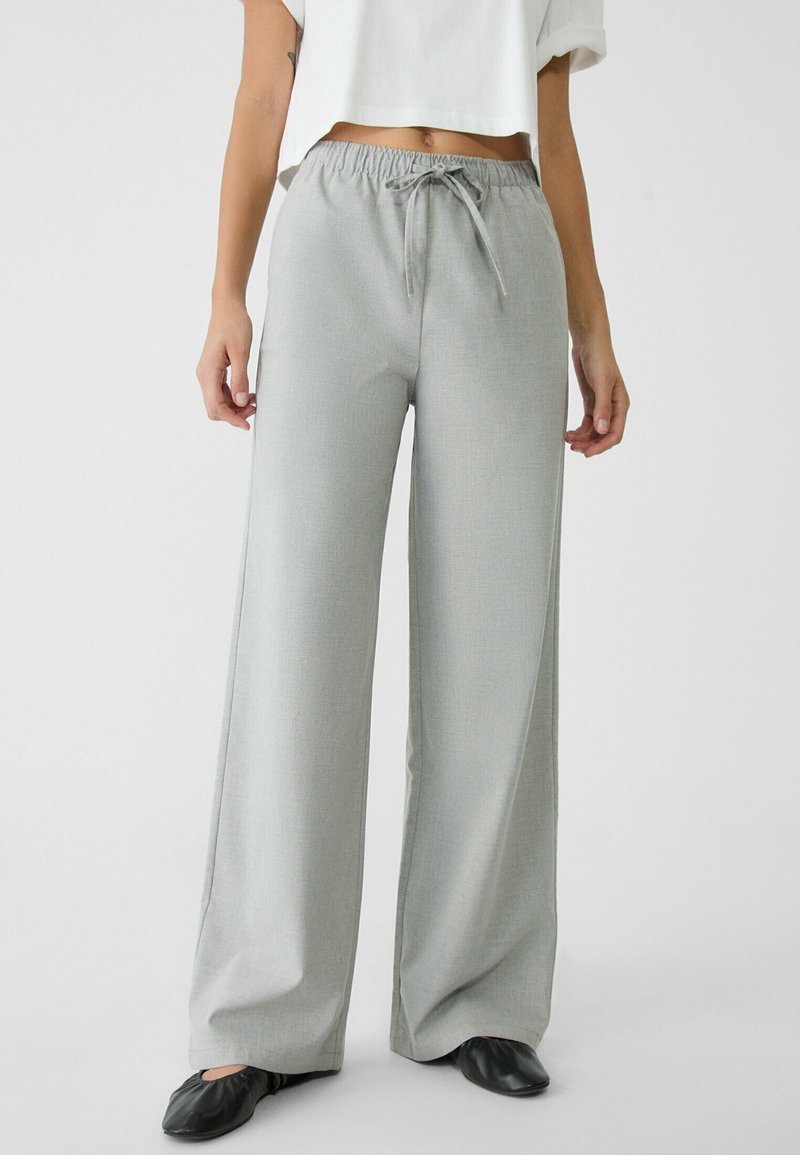 Kobieta ubrana w luźne jasnoszare spodnie z troczkami, krótki biały top i czarne baleriny, stojąca na tle jednolitej białej ściany.