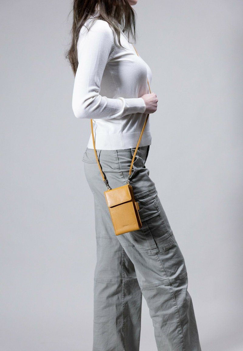 Mustard yellow leather crossbody bag with a rectangular shape, front flap, and adjustable strap, worn over gray cargo pants.