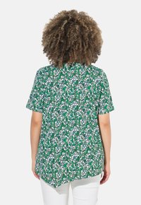 Blusa a maniche corte con un motivo floreale verde, caratterizzata da un orlo curvo e un tessuto testurizzato. Lo sfondo è bianco, che esalta la luminosità del design.
