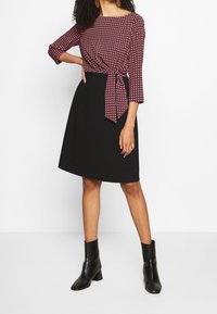 Blouse à motifs rouges et blancs avec une taille nouée, associée à une jupe noire évasée. Des bottines noires à la cheville complètent la tenue.