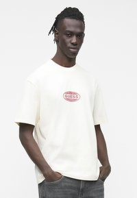 Camiseta de algodón color crema con mangas cortas, con un logo ovalado rojo de "GUESS" en el frente. La modelo tiene las manos en los bolsillos.