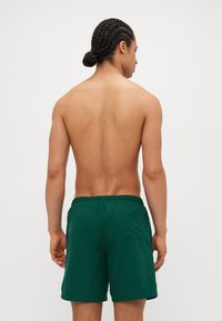 Pantaloni de baie verzi, cu un talie elastică, buzunar la spate și o textură netedă. Modelul este văzut de la spate, având torsul gol.